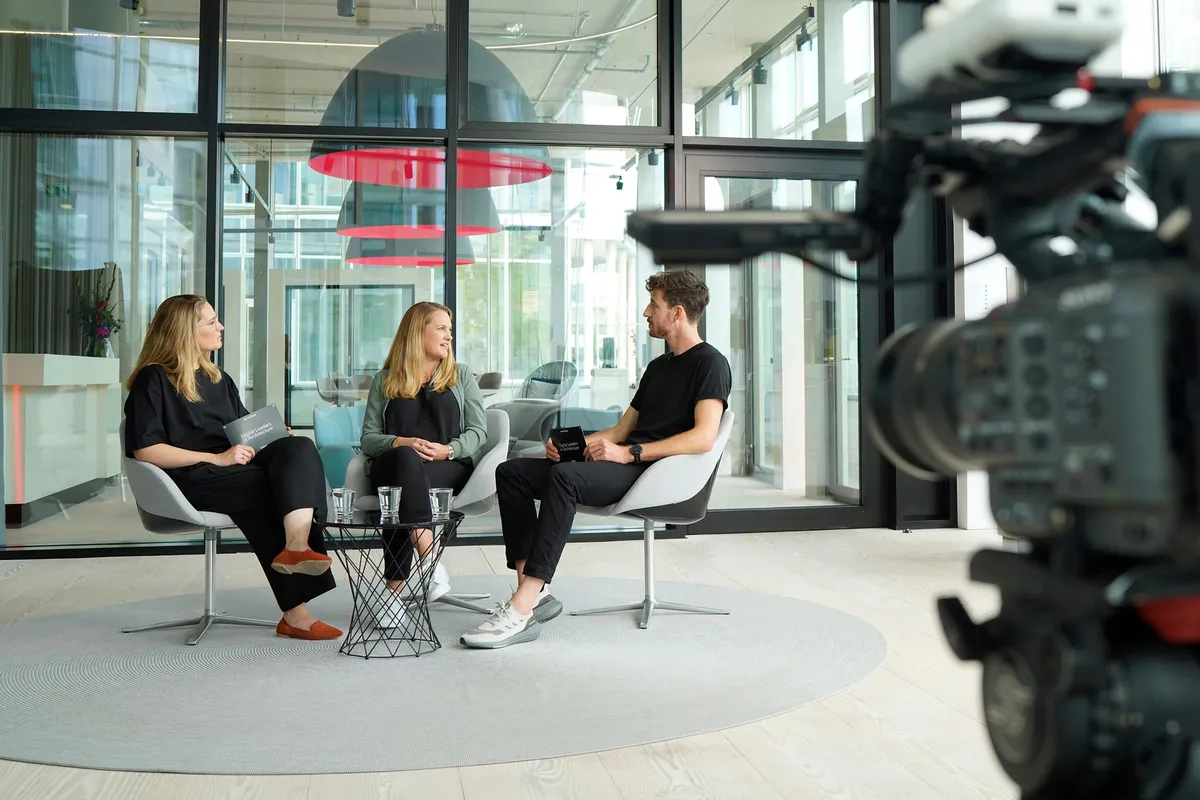 Two presenters and a guest sit on light-coloured swivel chairs in a light-filled Schüco showroom. A professional film camera can be seen in the foreground.