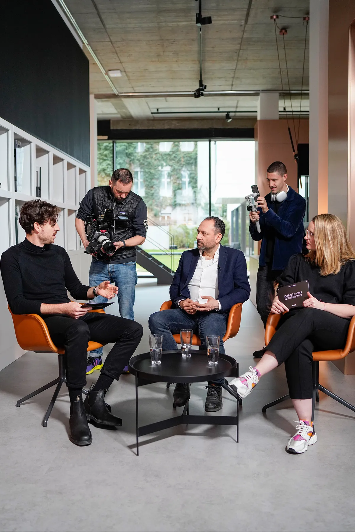 Drei Personen im Gespräch auf orangefarbenen Stühlen um einen schwarzen Tisch, zwei Kameraleute filmen die Runde für Digital Leaders in Architecture