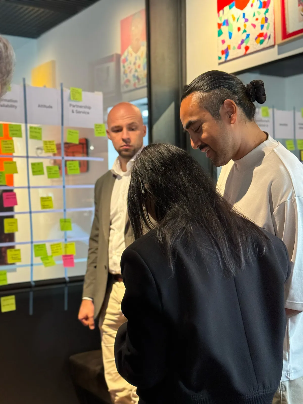 Drei Teilnehmende diskutieren vor einer Wand mit Haftnotizen, sortiert nach Themen wie »Partnership & Service«
