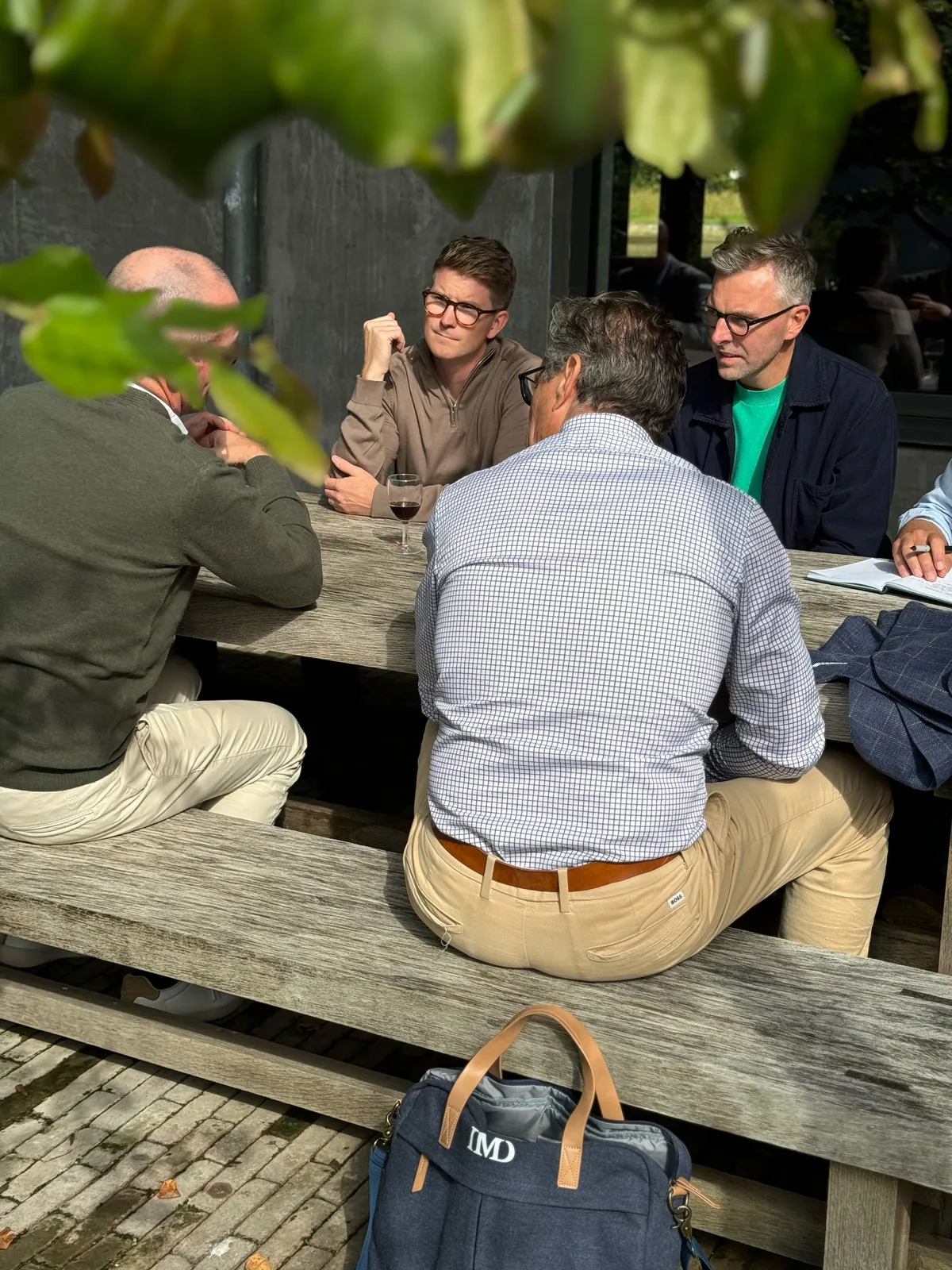 Teilnehmende im Gespräch an einem Holztisch im Freien unter Weinlaub