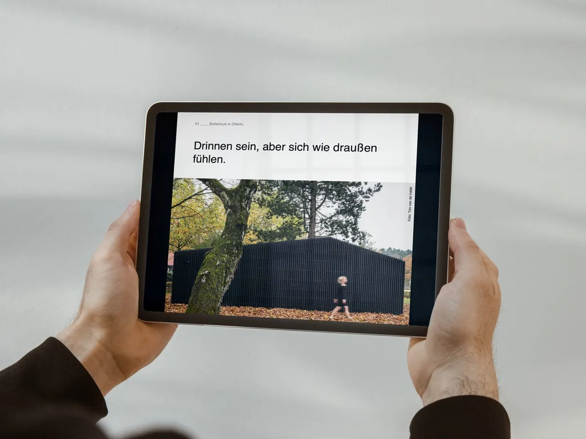 Hände halten ein Tablet mit einem digitalen Referenzbericht für Gira: Überschrift »Drinnen sein, aber sich wie draußen fühlen« über dem Foto eines dunklen Holzhauses zwischen Bäumen