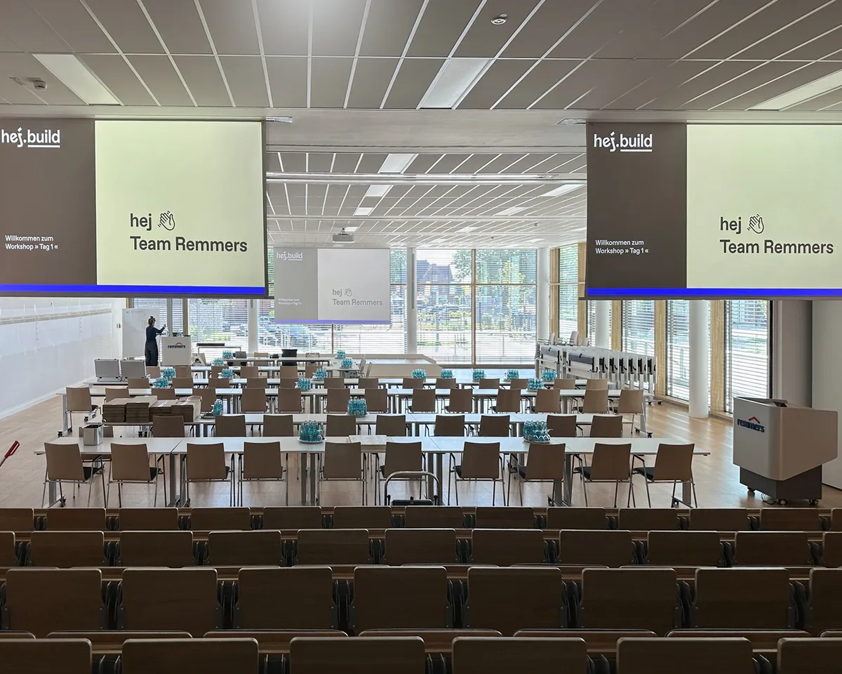 Seminarraum bei Remmers mit Tischreihen und zwei Leinwänden, auf denen hej.build das Team Remmers zum Workshop »Tag 1« begrüßt