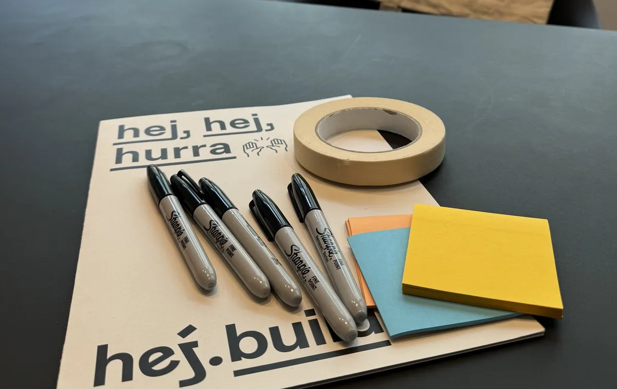 Workshop-Material auf einer hej.build-Mappe mit der Aufschrift »hej, hej, hurra«: Filzstifte, Kreppband und Haftnotizen