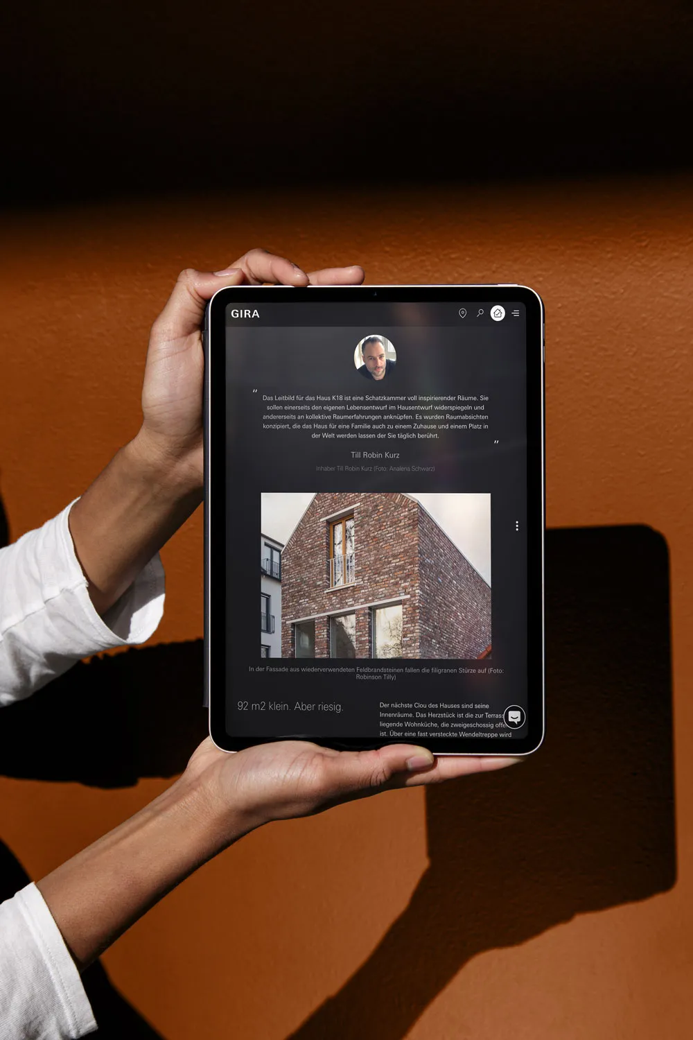 Hände halten ein Tablet mit einem Gira-Referenzbericht über ein Backsteinhaus vor orangebraunem Hintergrund