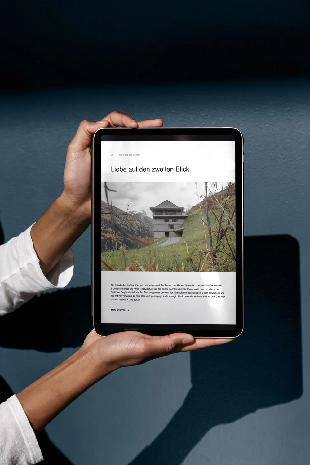 Hände halten ein Tablet mit dem Gira-Referenzbericht »Liebe auf den zweiten Blick« über ein Haus am Hang vor blauem Hintergrund