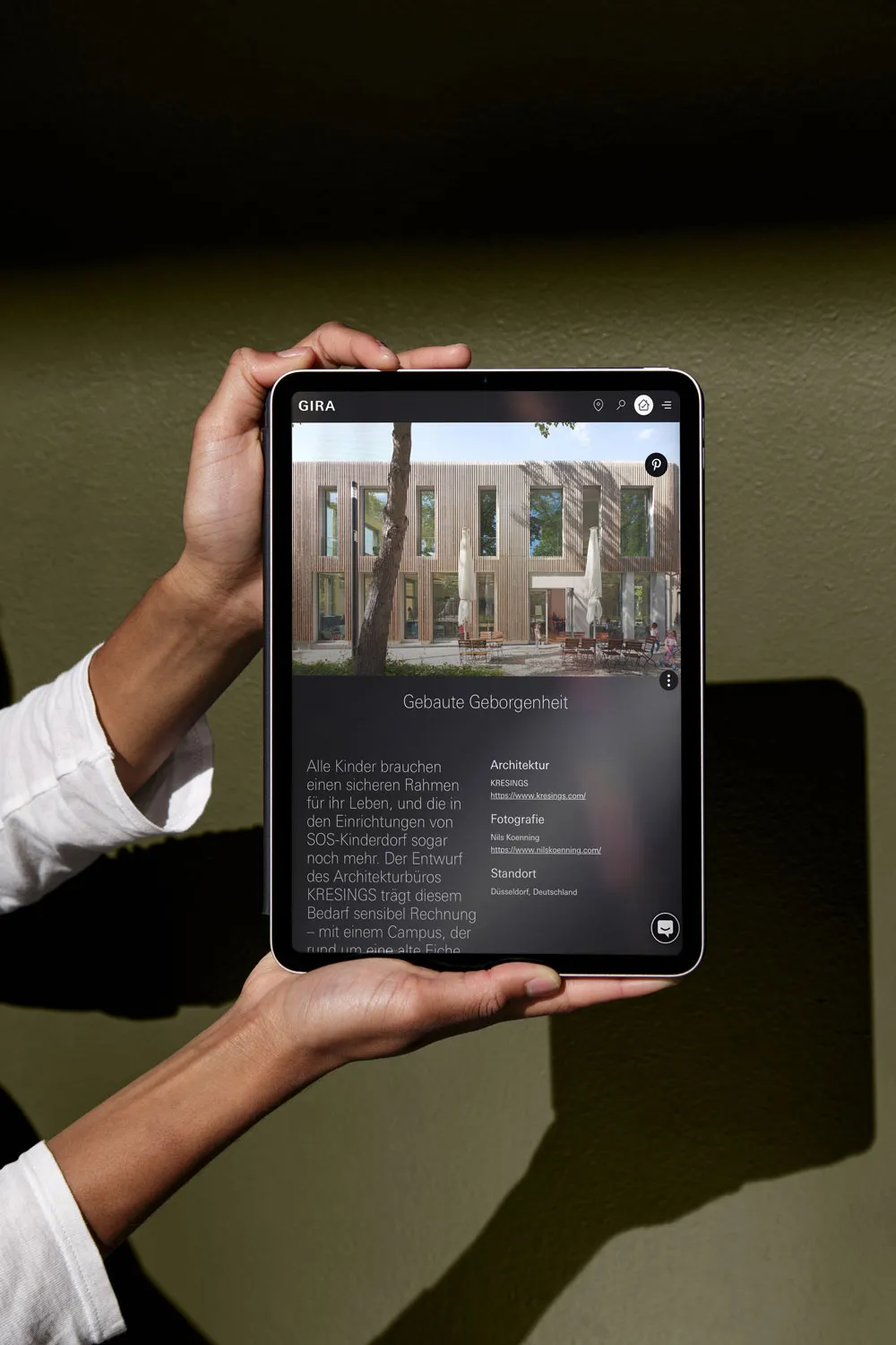 Hände halten ein Tablet mit dem Gira-Referenzbericht »Gebaute Geborgenheit« über ein SOS-Kinderdorf von KRESINGS vor olivgrünem Hintergrund