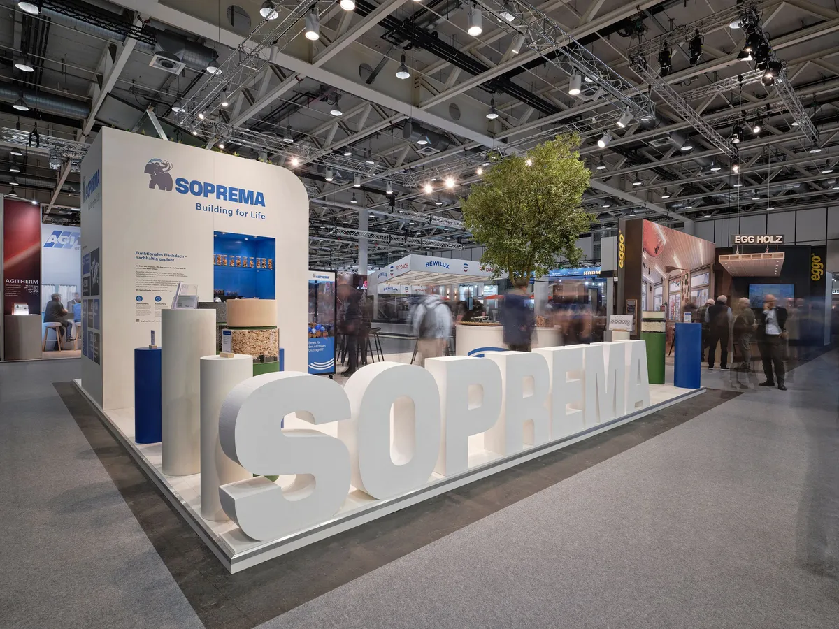 Soprema Messestand auf der Swissbau 2026 mit großem weißem Soprema-Schriftzug als Standkante, Baum und Infowand