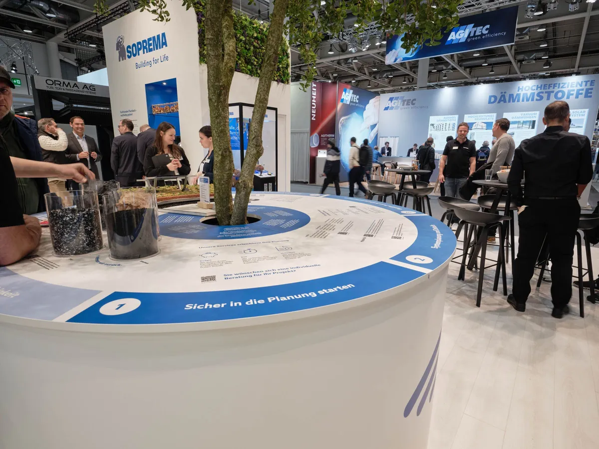 Runder Phasentisch am Soprema Messestand mit Materialproben in Gläsern und der Aufschrift »Sicher in die Planung starten«, dahinter Besucher:innen