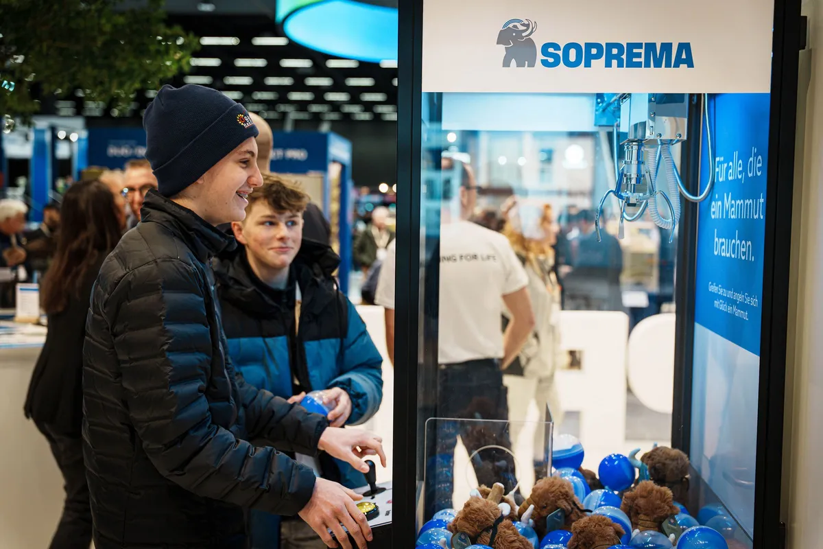 Zwei junge Besucher:innen spielen am Soprema Messestand an einem Greifautomaten mit Mammut-Plüschfiguren