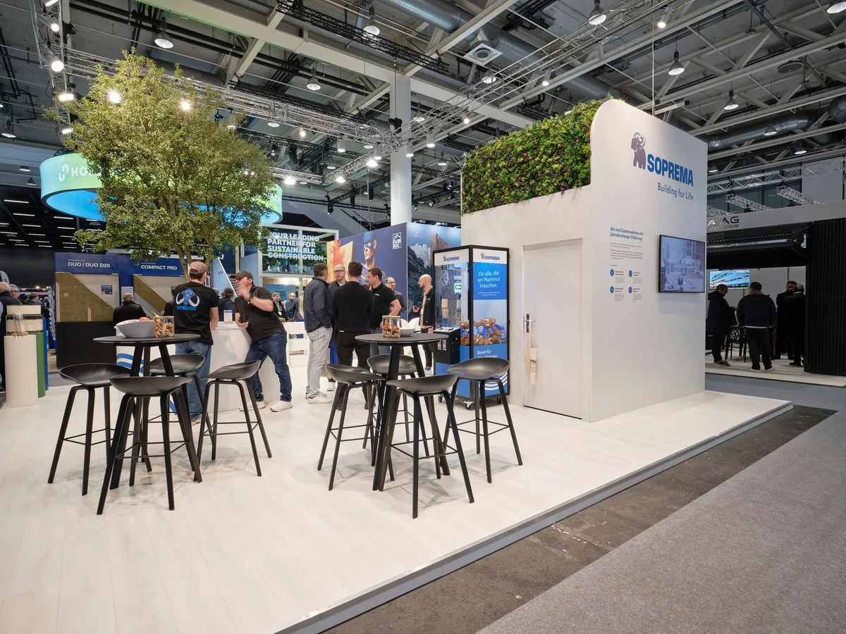 Lounge des Soprema Messestands mit Stehtischen, Barhockern und begrünter Standwand, Gäste im Gespräch