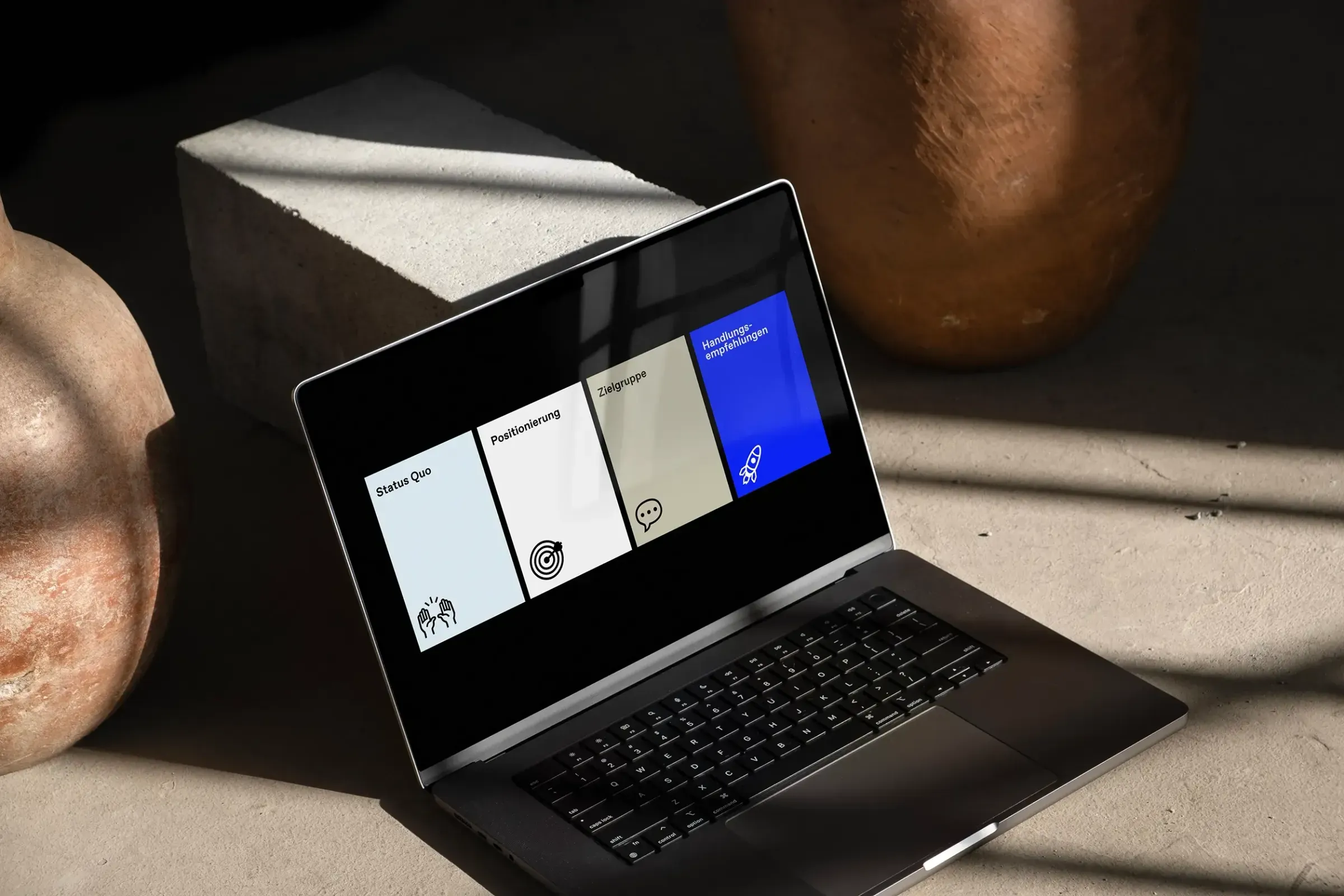 Laptop mit einer Präsentationsfolie in vier Feldern: Status Quo, Positionierung, Zielgruppe und Handlungsempfehlungen, daneben ein Betonblock und kupferfarbene Kugeln
