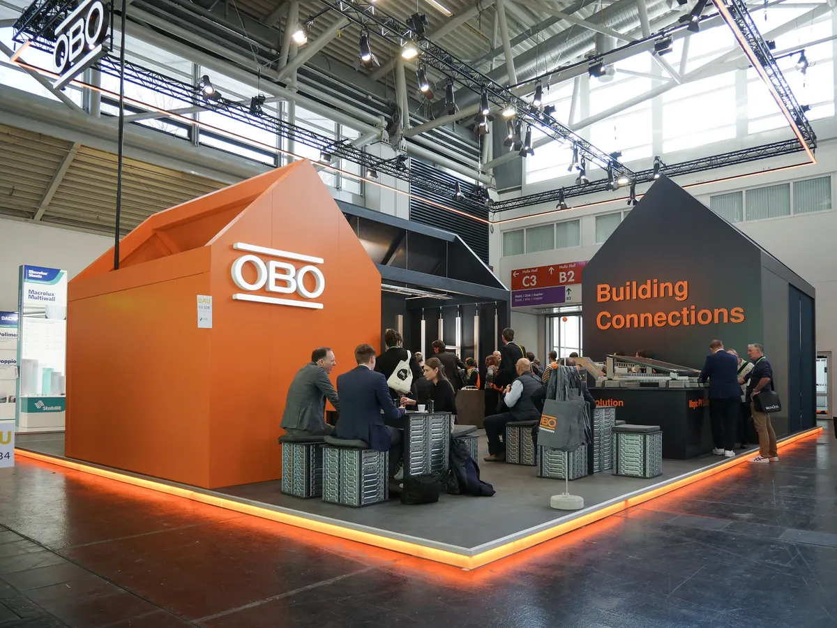 Orangefarbener hausförmiger Pavillon des OBO Messestands mit Logo und dem Schriftzug »Building Connections«, Gäste im Gespräch an Tischen