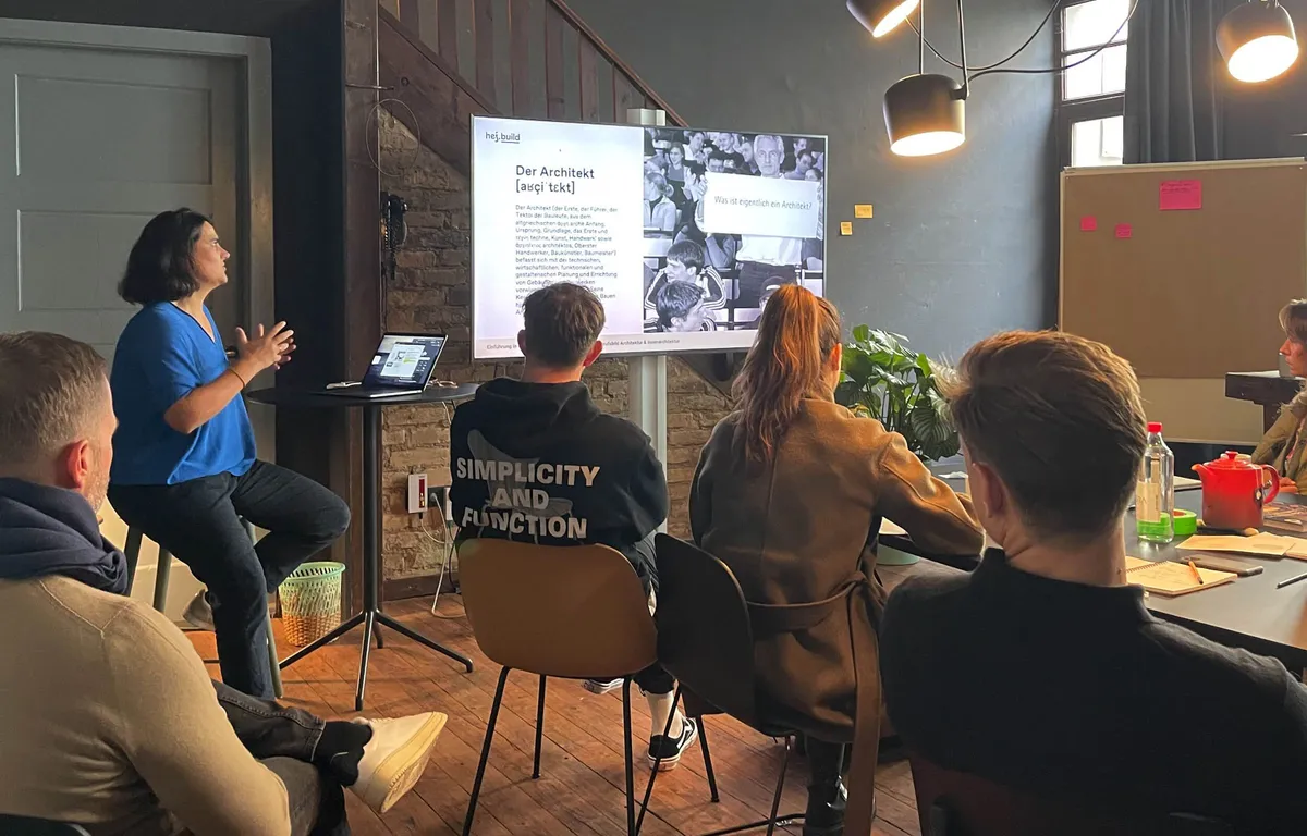 Präsentation beim Inhouse-Workshop für Muuto: Folie »Der Architekt« auf einem Bildschirm, Teilnehmende hören an Tischen zu