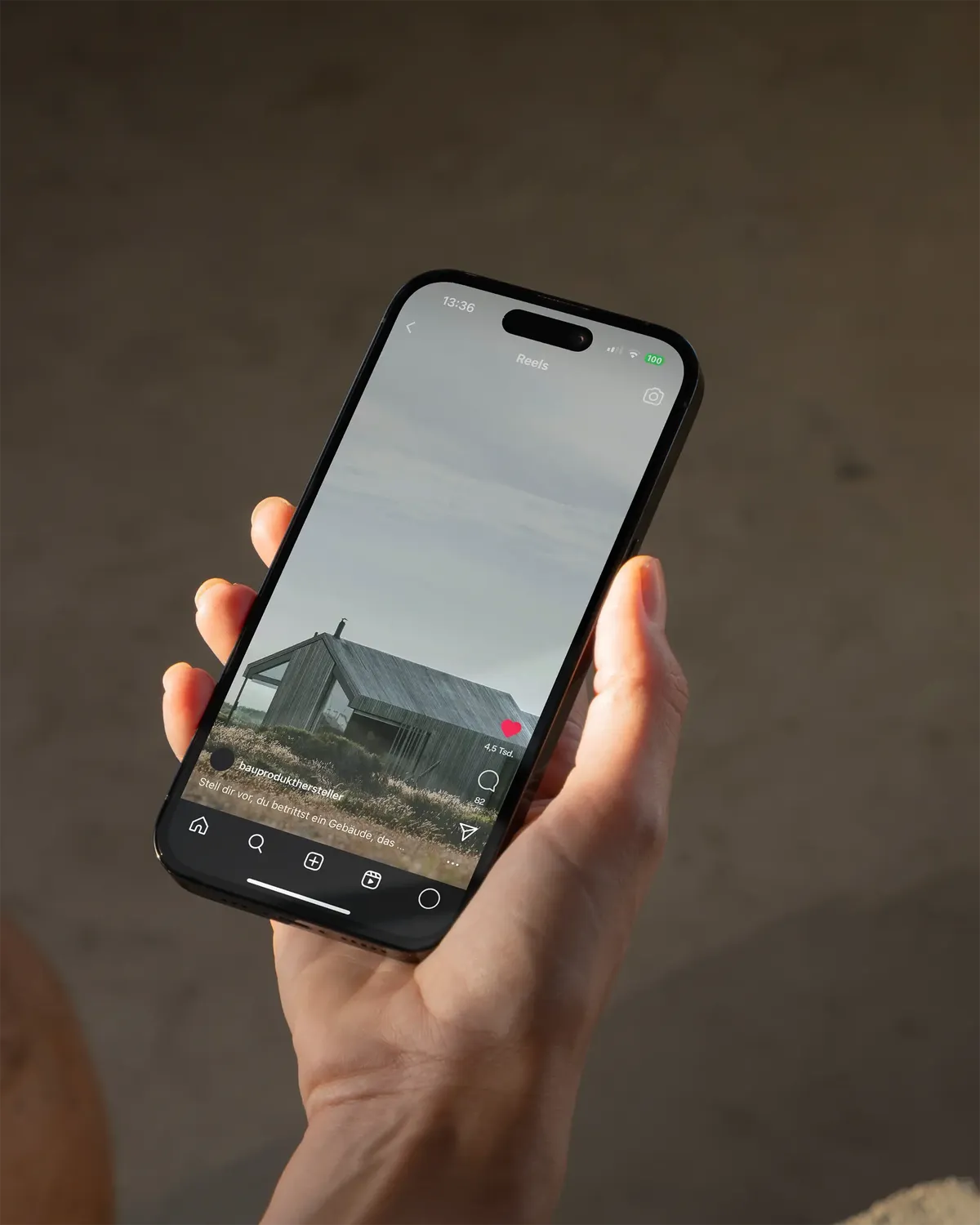 Eine Hand hält ein Smartphone mit einem Instagram-Reel, das ein modernes Holzhaus in einer ländlichen Landschaft zeigt