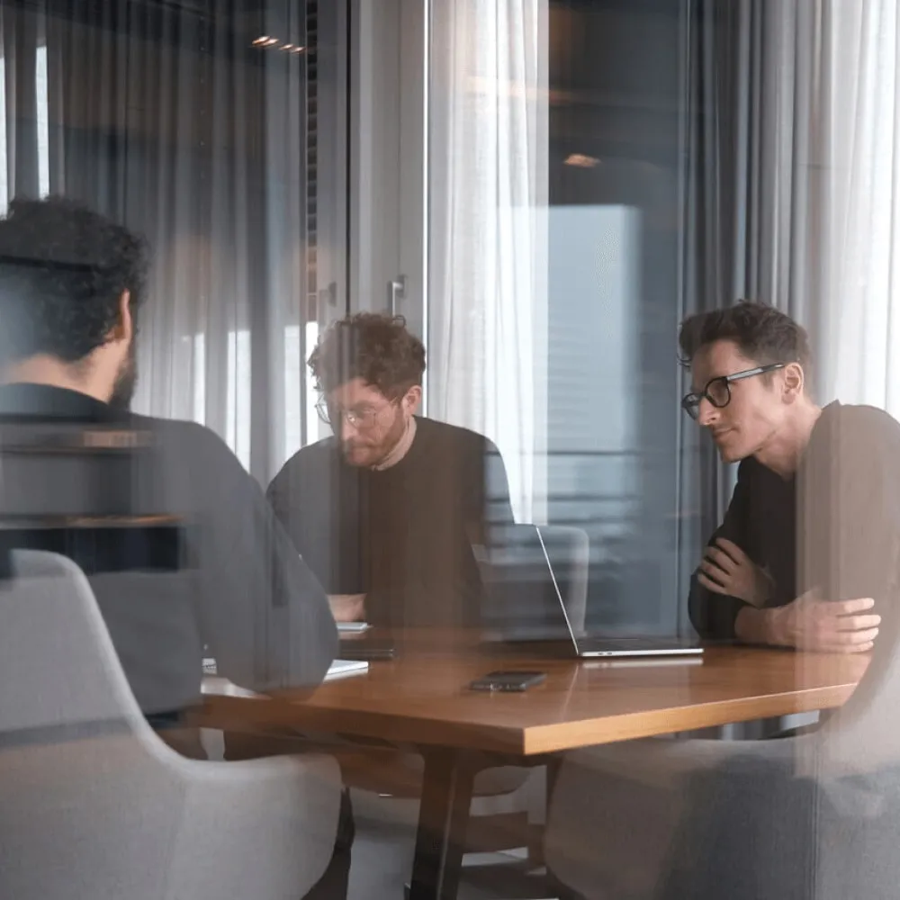 Drei Personen besprechen sich an einem Holztisch mit Laptop, fotografiert durch eine Glasscheibe