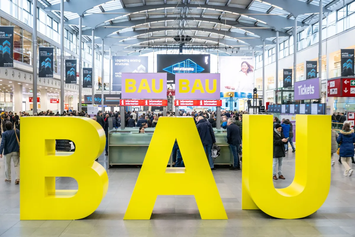 Große gelbe Buchstaben »BAU« in der Eingangshalle der Messe München, dahinter Besucher:innen an den Informationstheken und Schilder zur BAU.