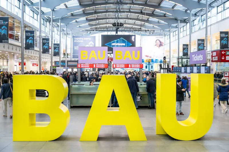 Große gelbe Buchstaben »BAU« in der Eingangshalle der Messe München, dahinter Besucher:innen an den Informationstheken und Schilder zur BAU.