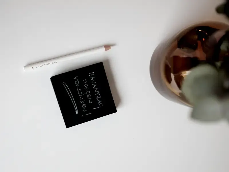 Schwarzer Post-it-Zettel mit handschriftlicher Notiz neben weißem Bleistift auf weißem Schreibtisch