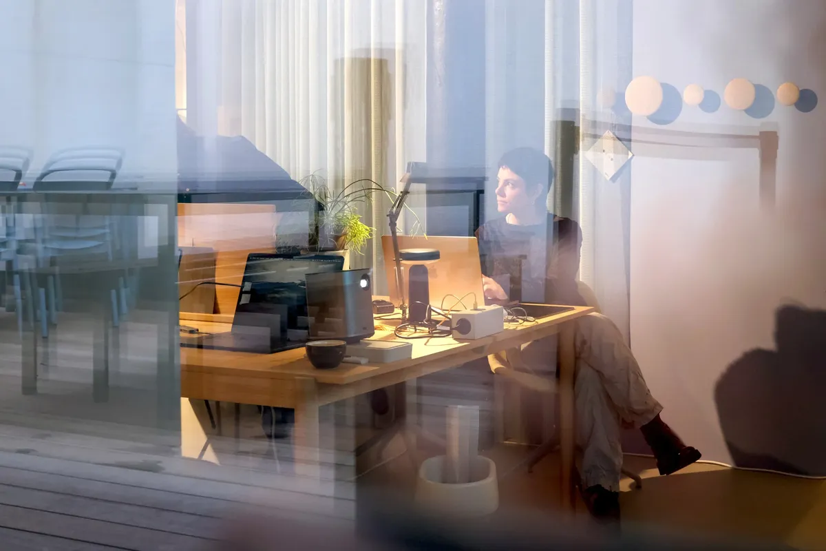 Person arbeitet an einem Schreibtisch am Fenster, im Vordergrund unscharfe Spiegelungen