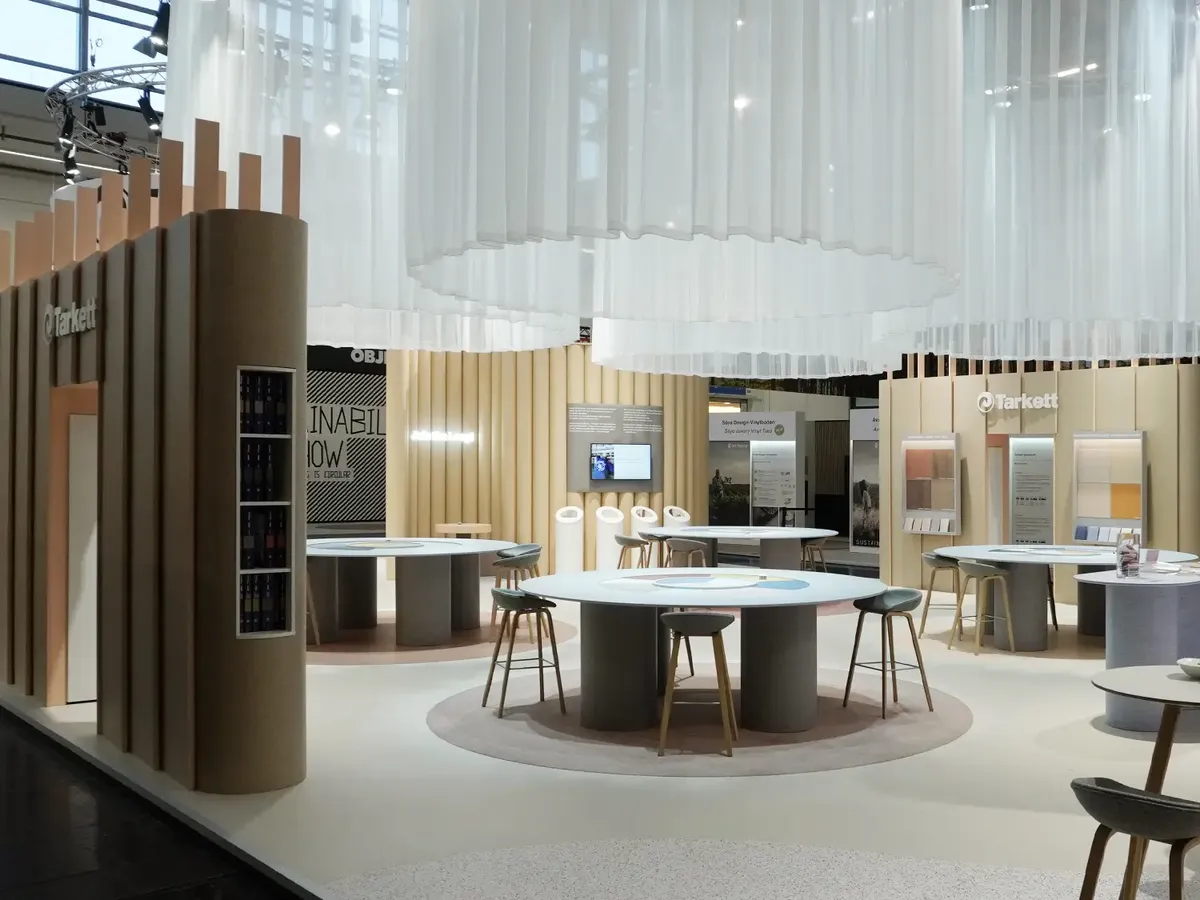 Tarkett Messestand mit weißer Stoffdeckeninstallation, runden Beratungstischen und cremefarbenen Lamellenwänden, präsentiert Bodenbelagslösungen auf der BAU 2025