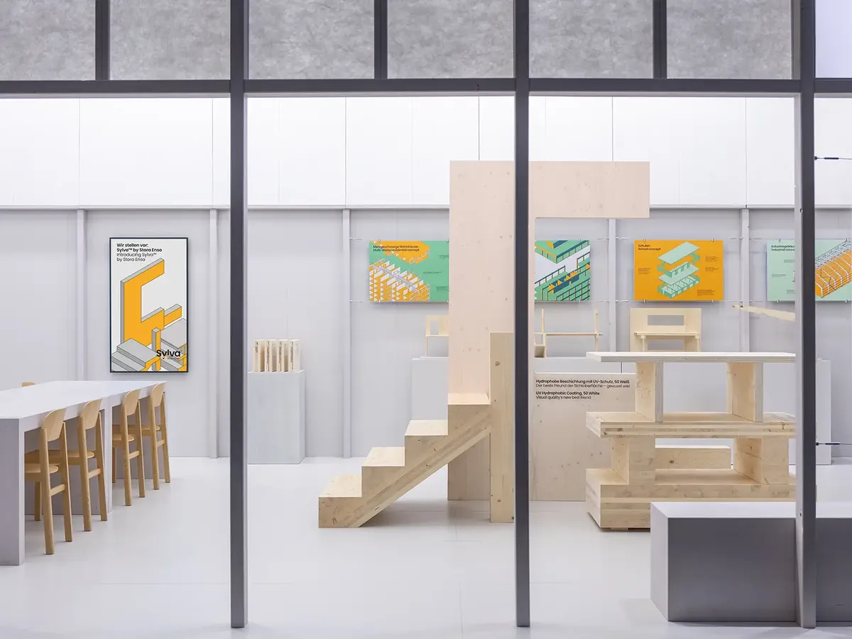 Stora Enso Messestand auf der BAU 2025 mit minimalistischem Design in Weiß und Holz, zeigt technische Illustrationen an der Wand und Holzkonstruktionen als Exponate, ergänzt durch einen Barbereich mit hellen Holzstühlen.