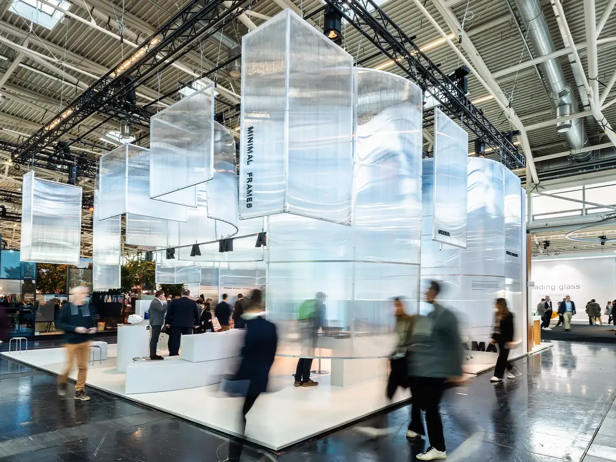 Messestand von Orama Minimal Frames auf der BAU 2025 mit schwebenden, transluzenten Paneelen über einer weißen Standfläche, davor Besucher:innen in Bewegung