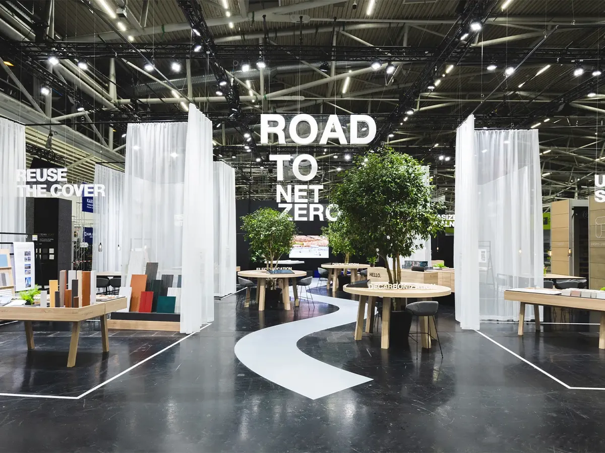 Agrob Buchtal Messestand mit geschwungener weißer Wegelinie auf dunklem Boden, die den 'Road to Net Zero' Claim visualisiert. Offene Architektur mit transparenten Vorhängen und Beratungszonen