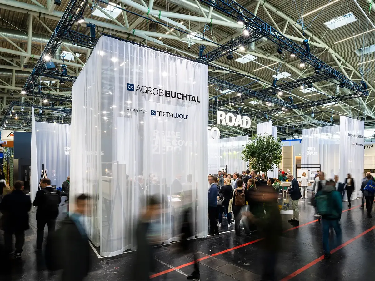 Agrob Buchtal Messestand mit transparenten Vorhängen und 'Road to Net Zero' Konzept, belebte Messeatmosphäre mit bewegten Besuchern im Vordergrund
