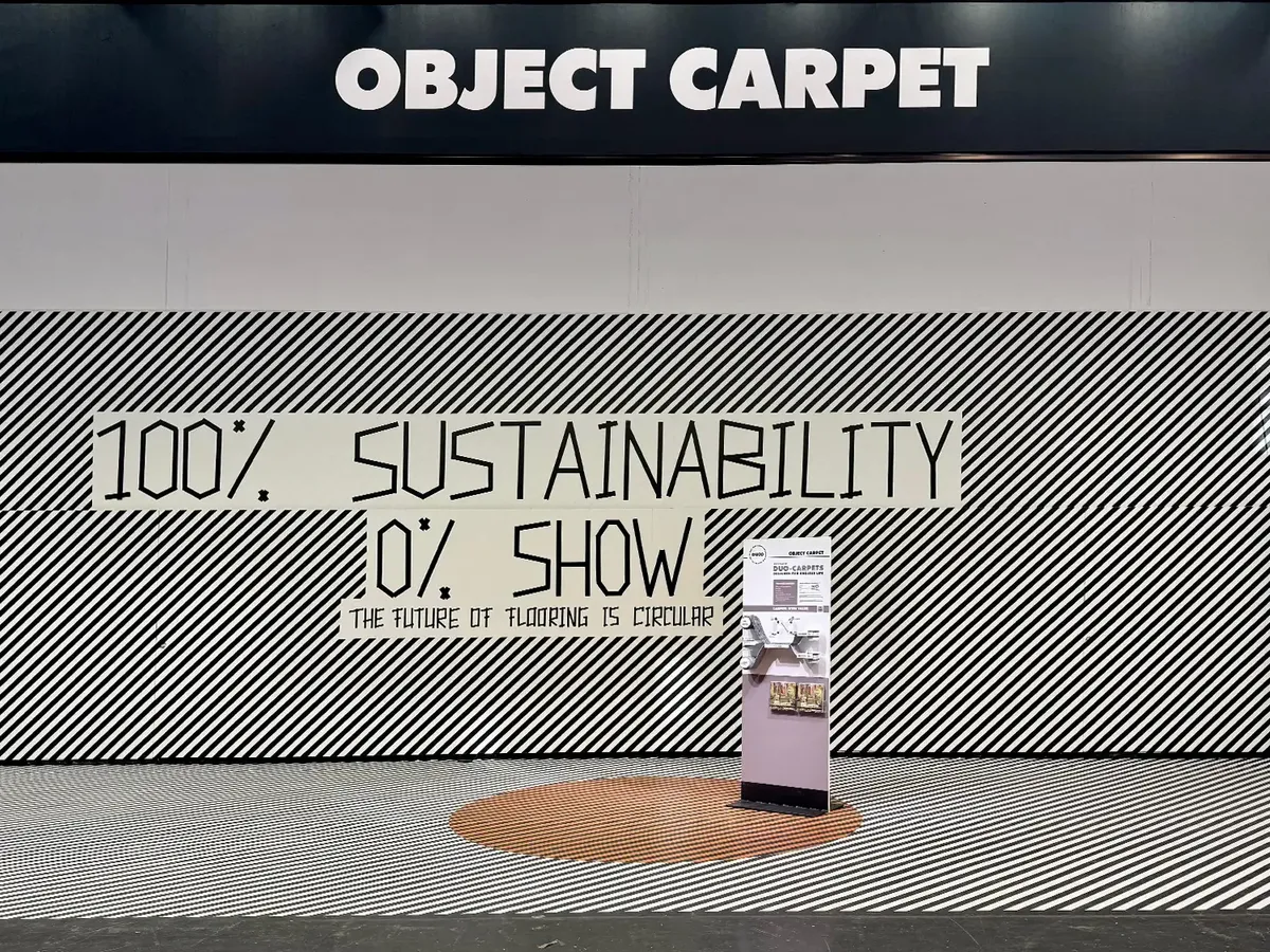 Object Carpet Messestand auf der BAU 2025 mit minimalistischem Konzept '100% Sustainability, 0% Show'. Schwarz-weiß gestreifte Wandgestaltung mit klarem Statement zur Nachhaltigkeit und einem einzelnen Produktdisplay auf orangefarbenem Kreisdetail.