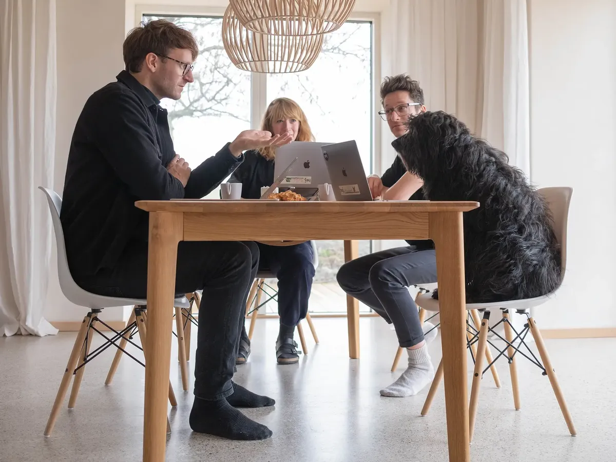 Das hej.build Führungsteam (Hannes Eberlein, Saskja Jagenteufel, Michael Rahmfeld und Bürohund Berta) in einer Teambesprechung am Holztisch, Ausdruck der kollaborativen und offenen Unternehmenskultur.