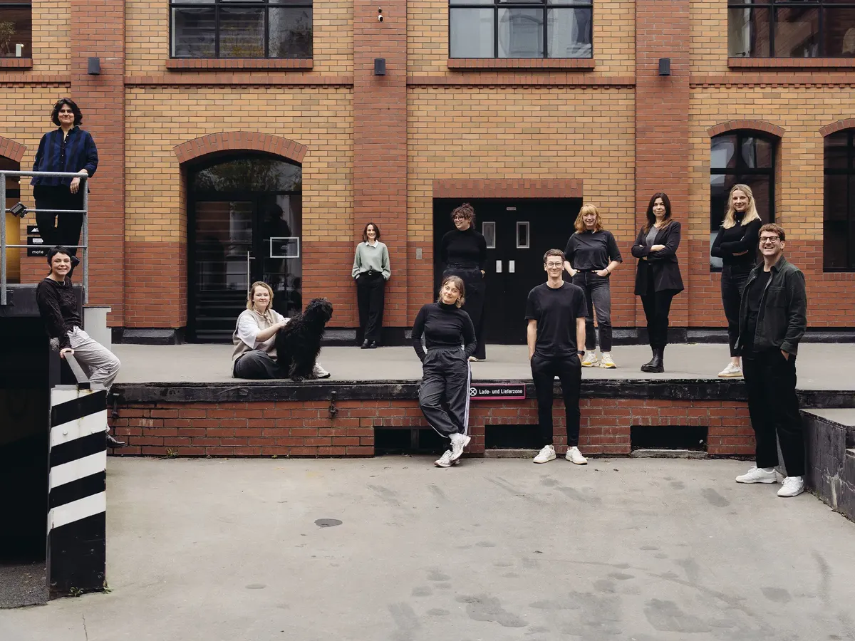 Das gesamte hej.build Team steht und sitzt lässig vor einer urbanen Backsteinmauer, Symbol der gewachsenen, vielseitigen und remote arbeitenden Strategieberatung.