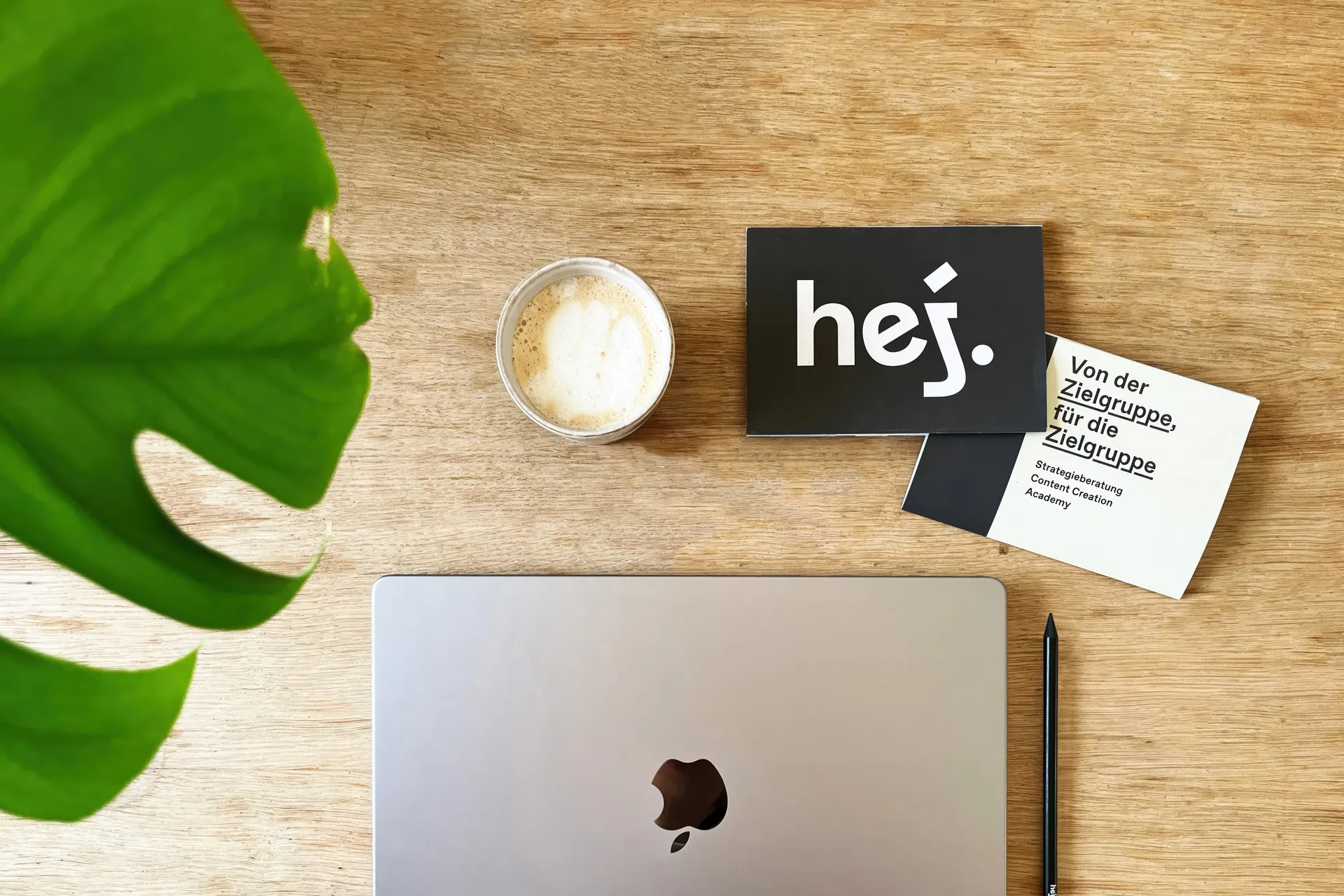 Holztisch von oben mit Kaffeetasse, Laptop, Stift, einem Monstera-Blatt und hej.build-Karten mit dem Satz »Von der Zielgruppe, für die Zielgruppe«
