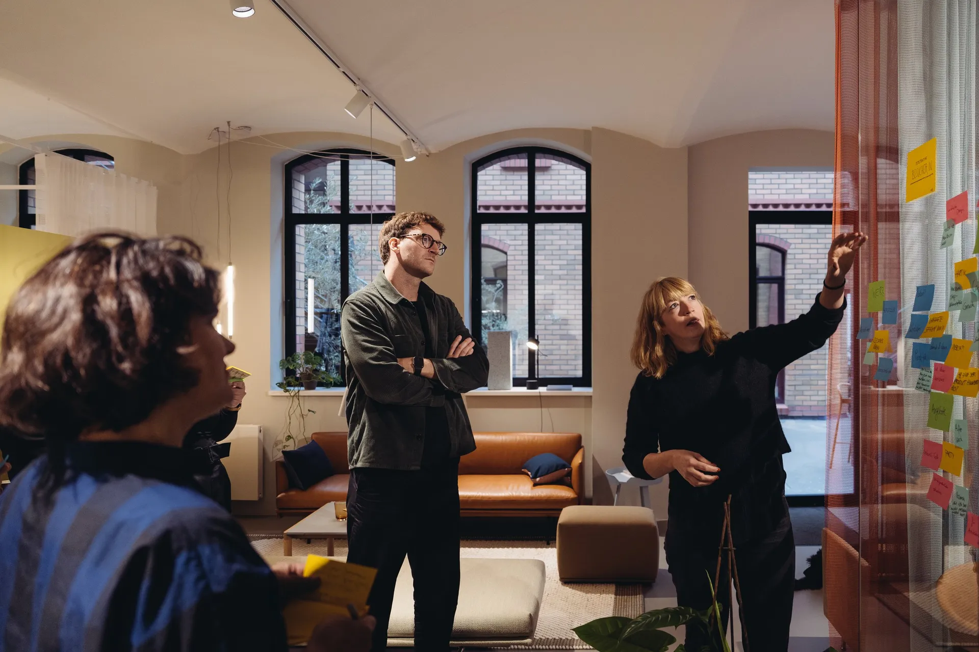 Workshop in einem Showroom: Eine Person zeigt auf Haftnotizen an einer Glaswand, zwei weitere hören zu