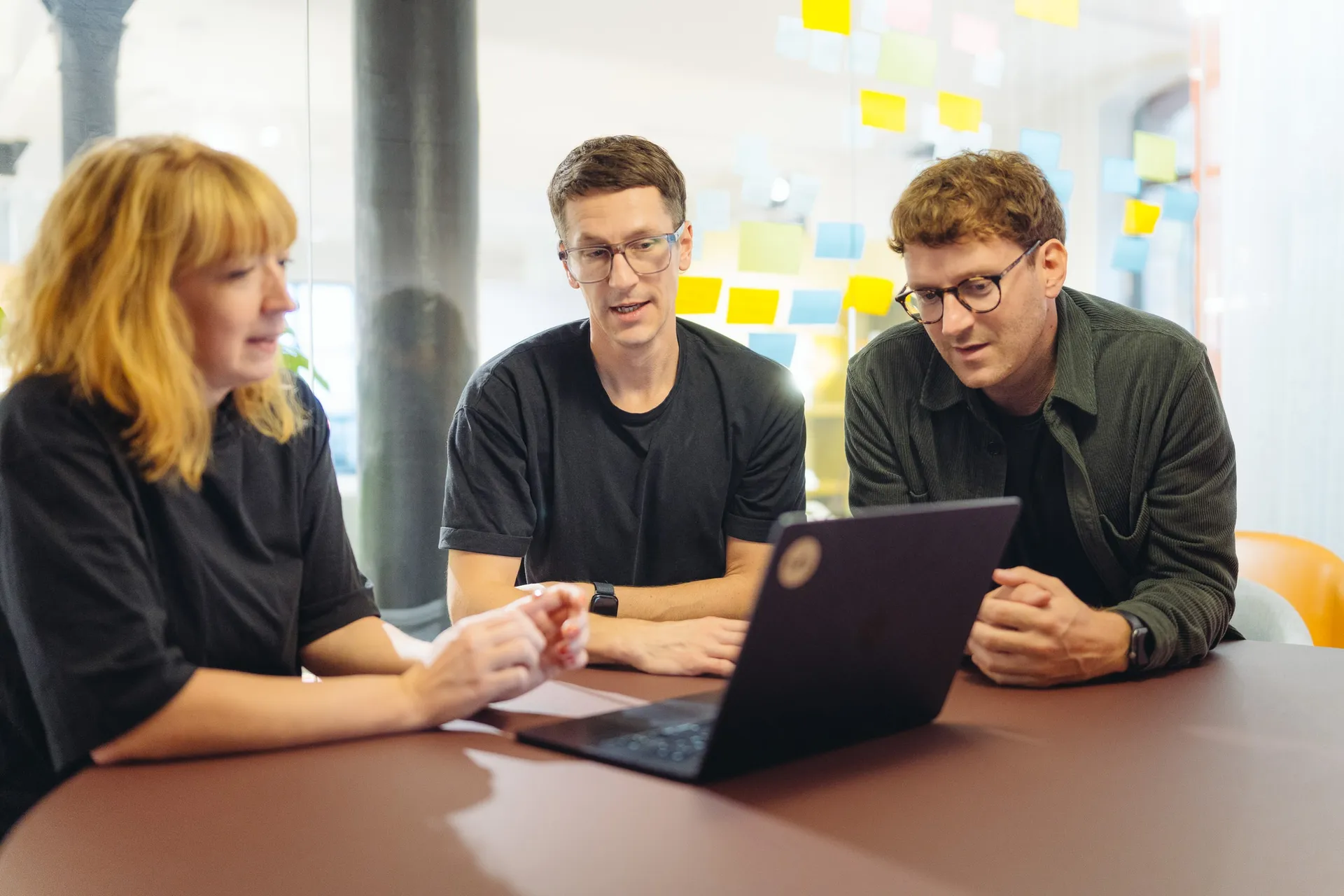 Drei aus dem Team im Gespräch am Laptop, dahinter eine Glaswand mit Haftnotizen