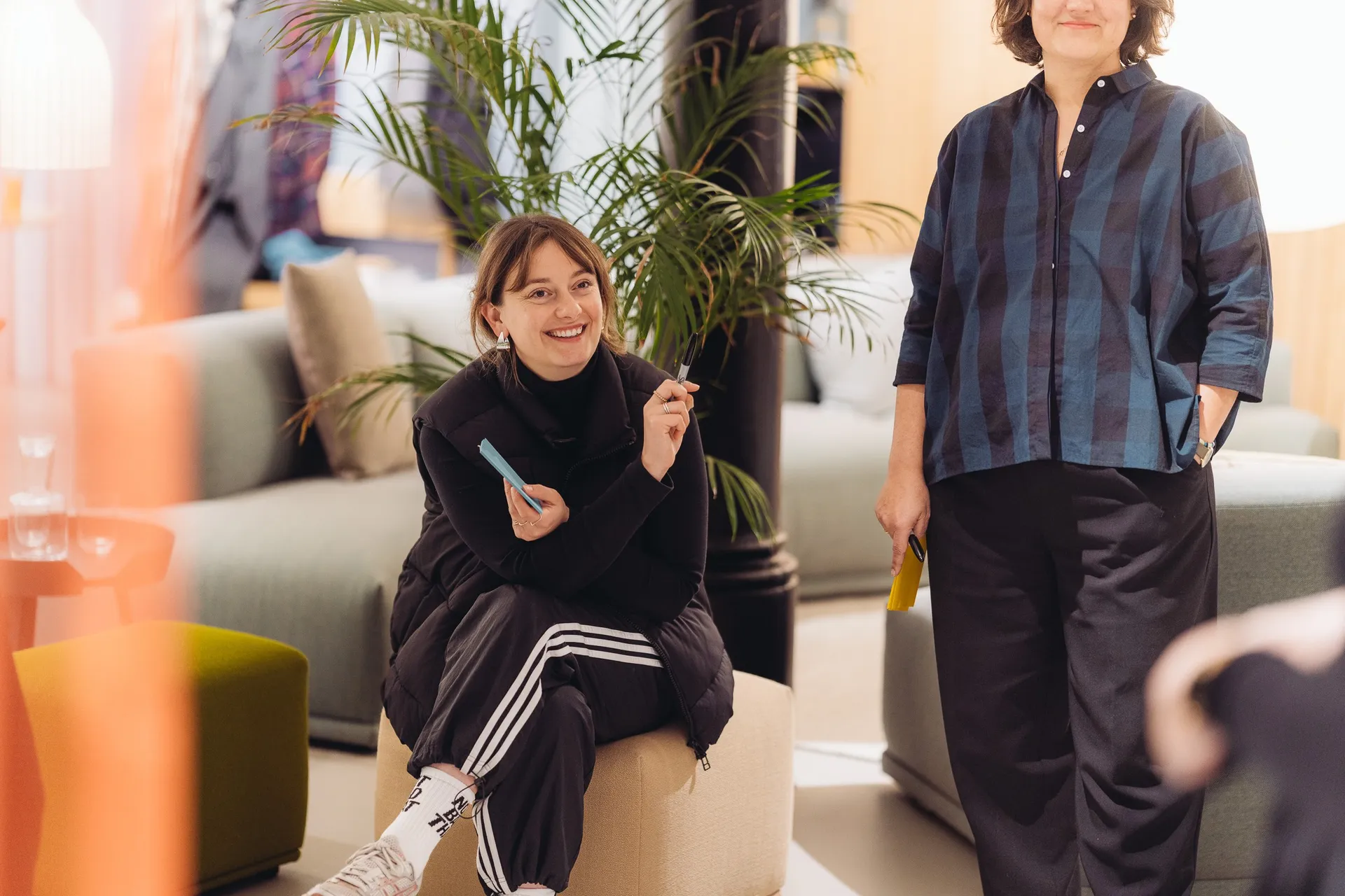 Eine Kollegin sitzt lachend mit Stift und Haftnotizen im Showroom, daneben steht eine zweite Person