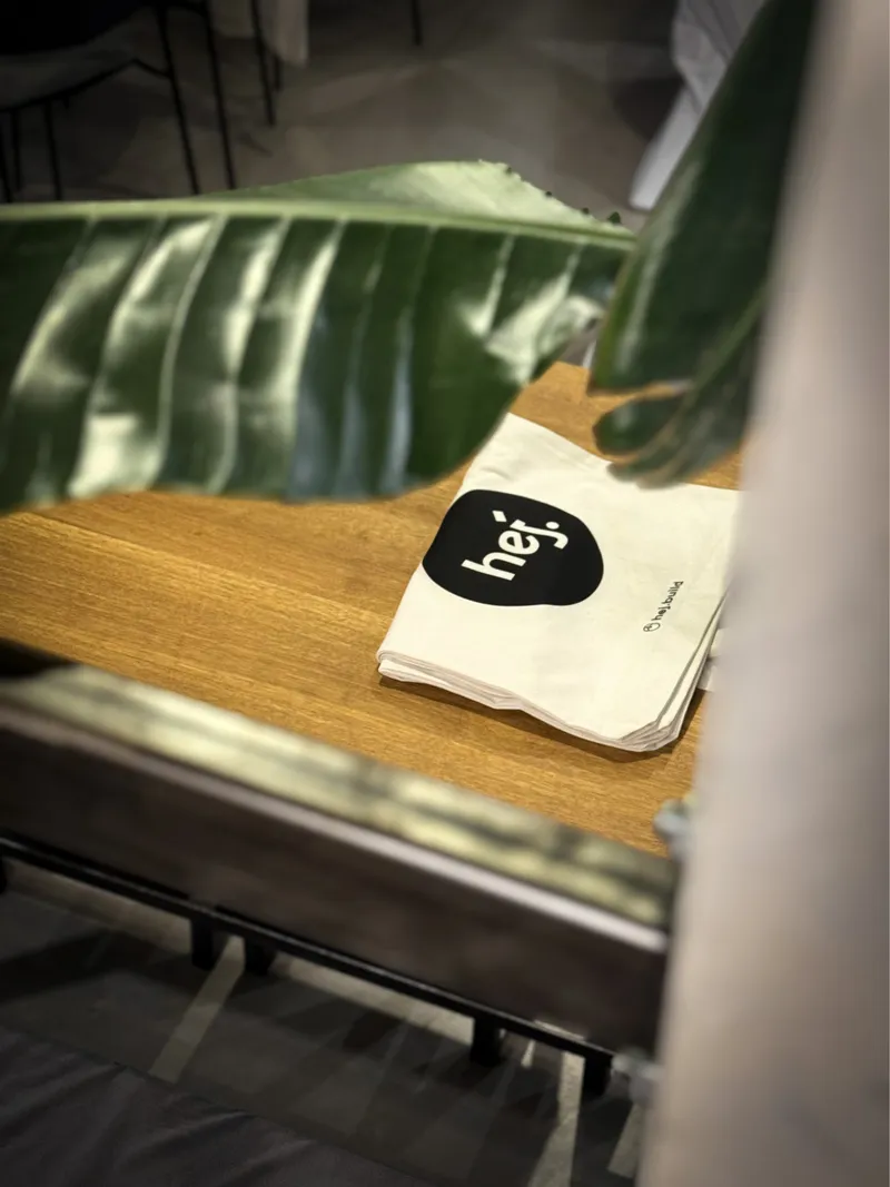 Stofftasche mit hej-Logo auf einem Holztisch, darüber ein großes Blatt